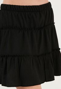 Jupe noire à volants en tissu texturé, avec un ourlet froncé et une taille élastique. Le tissu présente un léger motif froissé.
