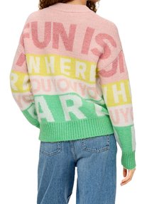 Person som bär en flerfärgad stickad tröja med stora bokstäver i rosa, gult och grönt, tillsammans med blå denimjeans, vänd bort.