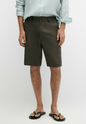 Man die donkerbruine knielange korte broek draagt, lichtblauw overhemd met lange mouwen met opgerolde mouwen en zwarte slippers.