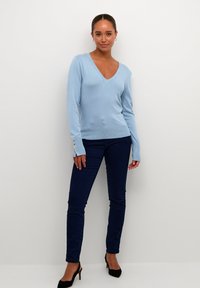 Lichtblauwe v-hals trui met lange mouwen, voorzien van gouden knoopaccenten op de mouwen, gecombineerd met donkere skinny jeans en zwarte hakken.