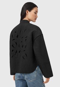 Donna con lunghi capelli scuri che indossa una giacca nera trapuntata con il logo "All Saints" ritagliato sulla schiena e jeans di denim blu.