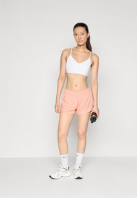 Bijela atletska bralette, ružičaste kratke hlače s elastičnim pojasom i bijele sportske cipele s crnim naglascima. Drži crnu bocu s vodom.
