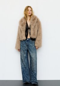 Jonge vrouw die een beige jas van nepbont draagt over een zwart cropped topje en wijde blauwe spijkerbroeken met witte verfspatten, staand tegen een witte muur.