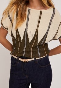 Kurzärmeliges, creme- und grün gestreiftes Oberteil mit strukturiertem Muster und dekorativen Akzenten, kombiniert mit dunklen Jeans und einem hellen Gürtel.