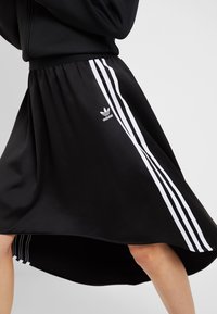 Svart träningskjol med vita tre ränder längs sidorna, en veckad elastisk midja och en flödande silhuett. Adidas-logga ingår.