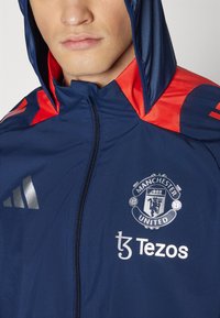 Chaqueta con capucha azul marino y detalles rojos. Presenta el logo de Adidas, el escudo del Manchester United y la marca Tezos, con cierre de cremallera completa.
