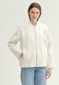 Weiße Zip-Jacke aus einem weichen, strukturierten Stoff. Verfügt über seitliche Taschen und eine entspannte Passform. Getragen über hellblauen Jeans.