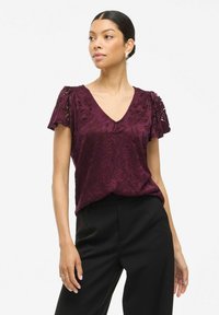Blusa in pizzo bordeaux con maniche a sbuffo corte, scollatura a V e motivo floreale, abbinata a pantaloni neri. Tessuto morbido e vestibilità rilassata.
