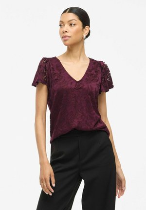 Blouse en dentelle bordeaux avec des manches bouffantes courtes, décolleté en V et motif floral, associée à un pantalon noir. Texture lisse et coupe décontractée.