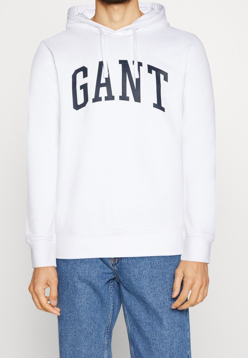 Sweat à capuche blanc en coton, avec une grande inscription noire "GANT" sur le devant, doté d'une capuche à cordon et de poignets côtelés. Associé à un jean bleu.