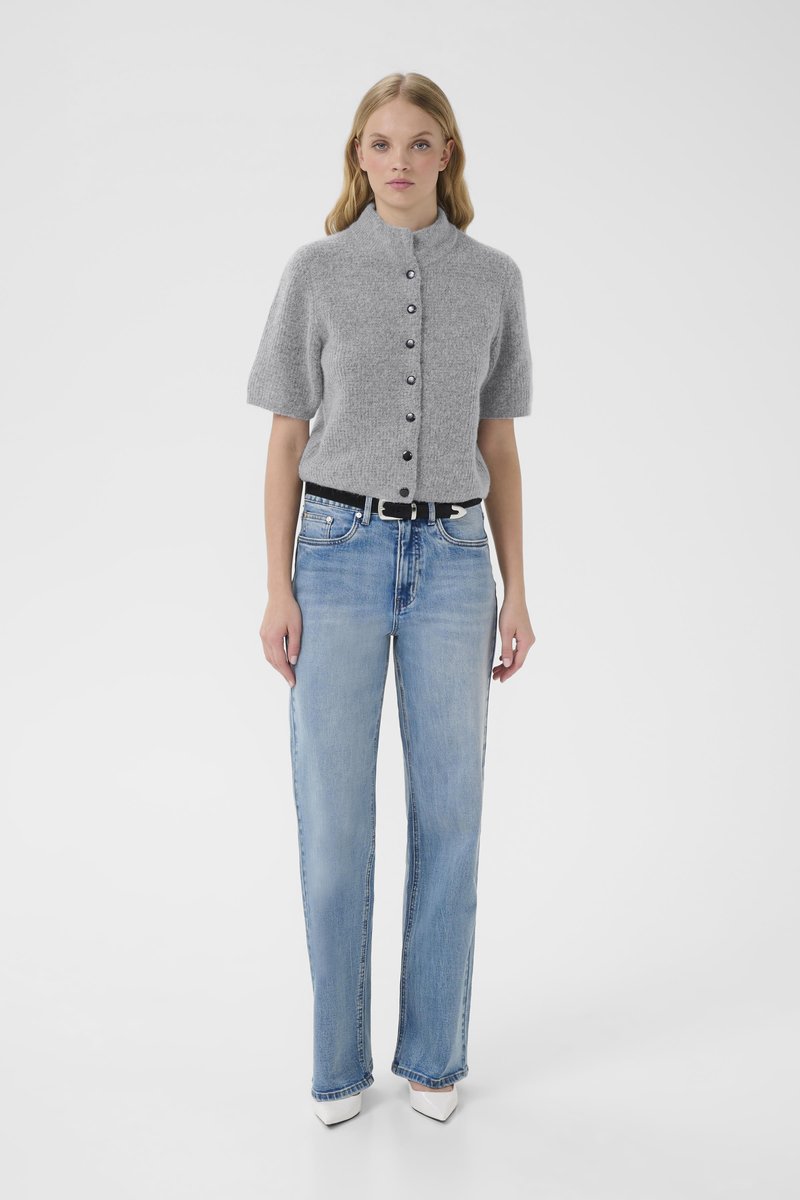 Woman standing wearing a grey short-sleeve buttoned sweater, light blue high-waisted jeans with a black belt, and white pointed shoes.