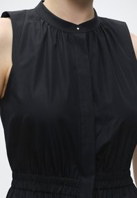 Gros plan d'une robe noire sans manches avec un col montant fermé par un petit bouton rond et une taille élastique froncée.
