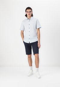 Chemise à manches courtes, bleu clair, avec des boutons foncés, associée à un short marine et des baskets blanches. Design simple, coupe décontractée, sans motifs.