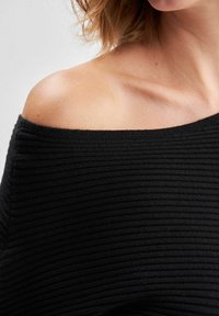 Haut noir épaules dénudées en tissu côtelé, présentant une texture horizontale et un large décolleté. Épaule et clavicule exposées visibles.