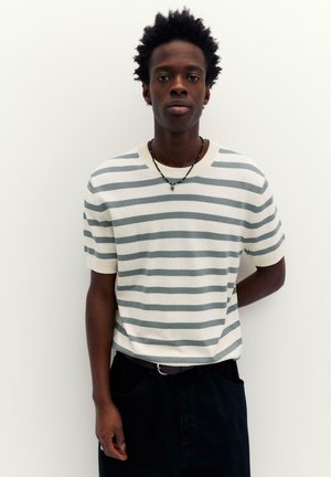 Mladý muž s krátkými kudrnatými vlasy, který má na sobě bílé a šedé pruhované tričko s krátkým rukávem a černé kalhoty, na jednobarevném bílém pozadí.