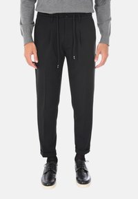 Pantaloni neri su misura realizzati in tessuto liscio, con vita regolabile con cordoncino, pieghe frontali e orli arrotolati, abbinati a scarpe nere.