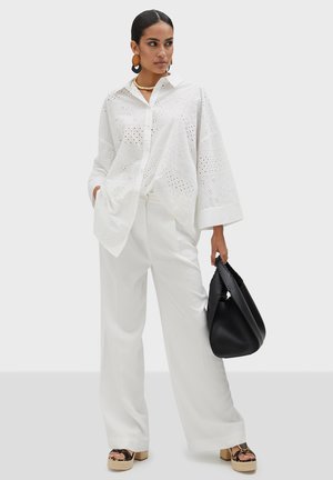 Blouse blanche en broderie anglaise à manches longues, associée à un pantalon blanc à jambes larges. Le mannequin tient un sac à main noir texturé et porte des sandales à plateforme.