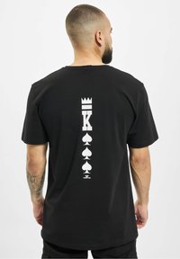 Black cotton t-shirt featuring a large white graphic design on the back, including a crown, letter "K," and spade symbols.