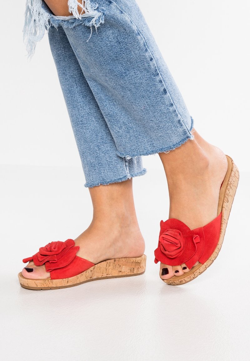 Pieds portant des sandales compensées rouges à semelles en liège avec une grande décoration florale en tissu, assorties à un jean bleu clair déchiré coupé au-dessus de la cheville.