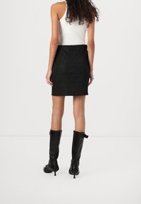 Femme portant un haut blanc sans manches, une mini-jupe noire texturée et des bottes noires à talons montant jusqu’aux genoux, debout devant un fond clair uni.