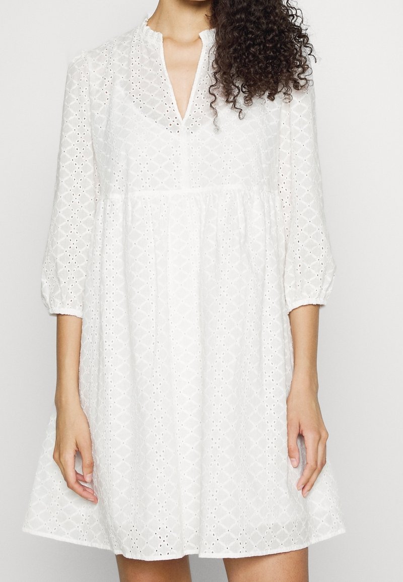 Femme portant une robe blanche en broderie anglaise, manches trois-quarts et décolleté en V, avec des cheveux bouclés tombant sur une épaule.