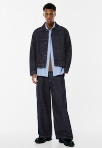 Crna traper jakna s kutijastim krojem, uparena s svijetloplavom košuljom s ovratnikom i širokim tamnoplastičnim hlačama. Crne masivne čizme zaokružuju izgled.