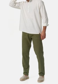 Biała koszula z długim rękawem z kołnierzykiem typu henley zapinanym na guziki, zestawiona z oliwkowozielonymi spodniami sztruksowymi z pionowymi paskami oraz szarymi wsuwanymi butami.