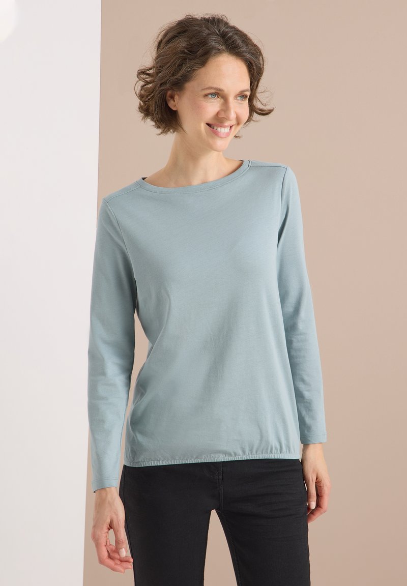 Light blue long-sleeve shirt made of soft fabric, featuring a round neckline and a slightly gathered hem, paired with black trousers.