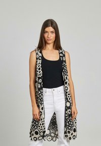 Ärmellose schwarze und cremefarbene gehäkelte Weste mit floralen Mustern und offenem Design, kombiniert mit einem schwarzen Oberteil und weißen Jeans.