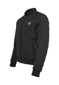 Veste bomber matelassée noire avec col, poignets et ourlet côtelés, fermeture éclair frontale, poches latérales et petite pièce en cuir sur la poitrine.