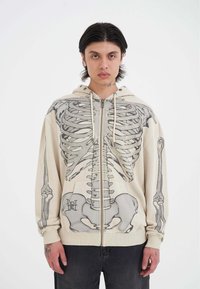 Felpa con zip in crema con stampa grafica di uno scheletro grigio, dotata di tasca a marsupio e cappuccio con coulisse. Composizione in cotone misto, vestibilità comoda.