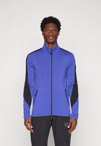 Zila jaku ar melniem akcentiem, garām piedurknēm un divām rāvējsl�ēdzēju kabatām. Gluda tekstūra, sportiska piegriezuma un augstas apkakles dizains.