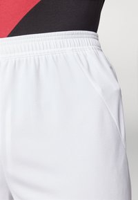 Vita sportshorts med elastisk midja, strukturerad väv, sidofickor och en jämn yta. Närbild som lyfter fram materialet.