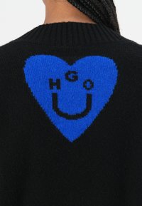 Black knit sweater featuring a large blue heart design on the back, with the letters "HGO" and a smiley face pattern integrated.