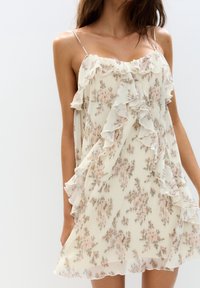 Vestido sin mangas en color crema con estampado floral y acentos de volantes. La tela ligera presenta tonos suaves de rosa y verde, creando una silueta fluida.