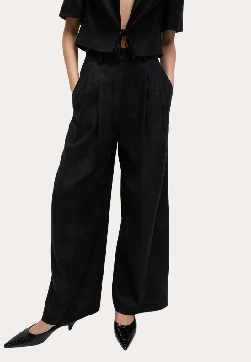 Femme portant un pantalon noir taille haute à jambes larges avec plis à l'avant, associé à des escarpins noirs à bout pointu et un top court noir noué.