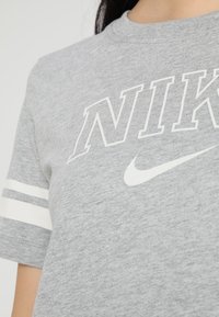 Camiseta gris de Nike con texto y logo "NIKE" en blanco contorneado, que presenta dos rayas blancas en la manga izquierda.