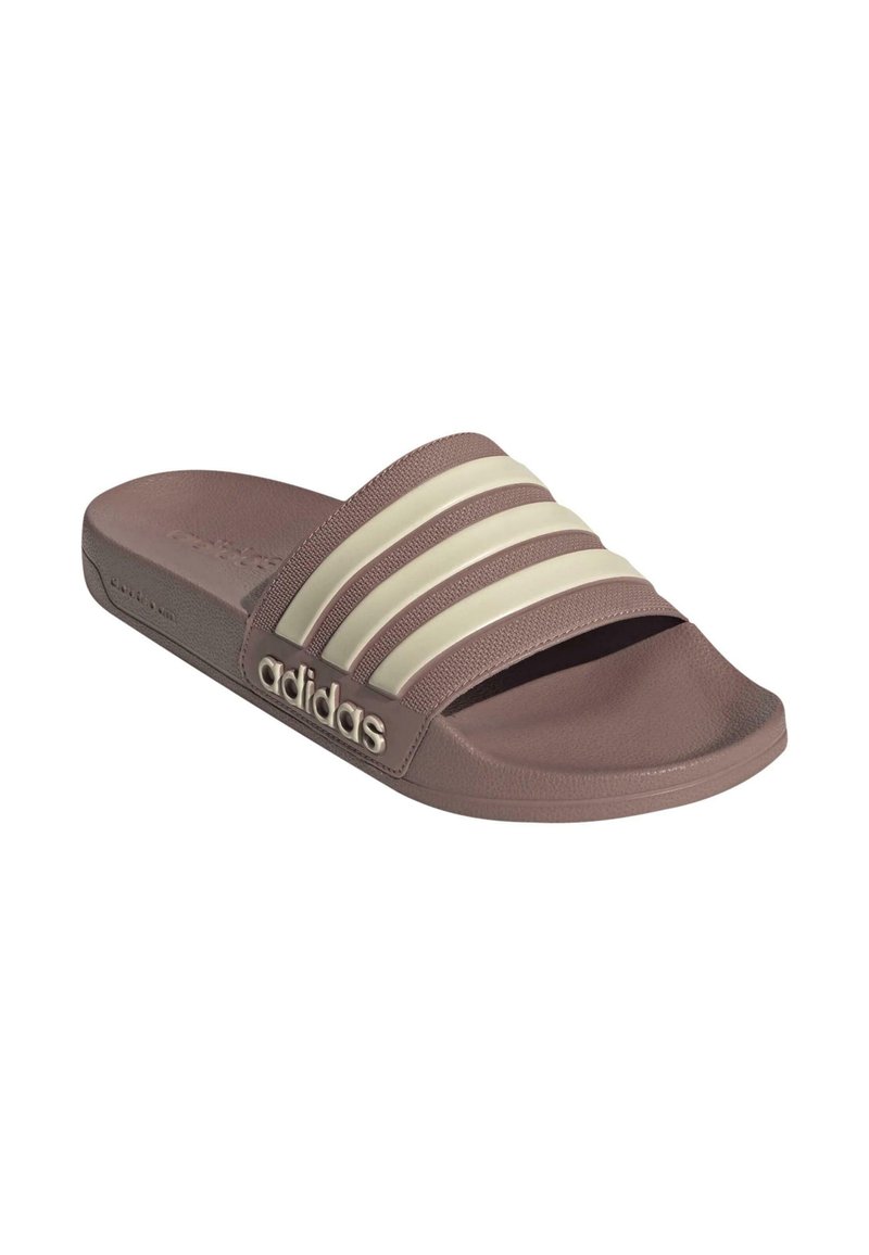 Braune Slipperschuhe mit drei cremefarbenen Streifen und strukturiertem Fußbett. Mit Adidas-Logo an der Seite und einer flachen, gepolsterten Sohle.