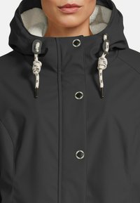 Vrouw in een zwarte gewatteerde jas met een capuchon, voorzien van een witte fleece voering en witte koordjes met zwarte uiteinden en knopen.