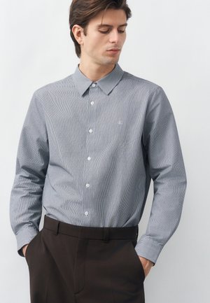 Homme portant une chemise grise claire à boutons avec un petit motif à carreaux et un pantalon marron foncé, debout les mains dans les poches.