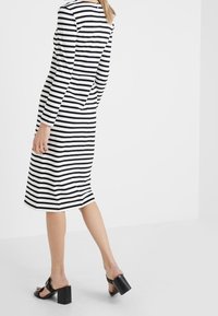 Kobieta w midi sukience w poziome czarne i białe paski, w czarnych sandałach na obcasie, stojąca na tle jednolitego białego tła.