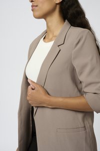 Femme portant un blazer beige sur un haut blanc, ajustant le blazer avec la main gauche, visible des épaules à la taille.