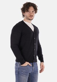 Cardigan negro hecho de suave tejido de punto, con escote en V, mangas largas y seis botones en la parte delantera. Llevado sobre una camiseta blanca.