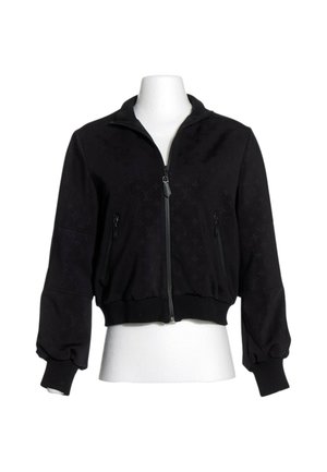 Veste noire zippée, manches longues, poignets et bas côtelés, avec deux poches avant zippées, présentée sur un torse de mannequin blanc.