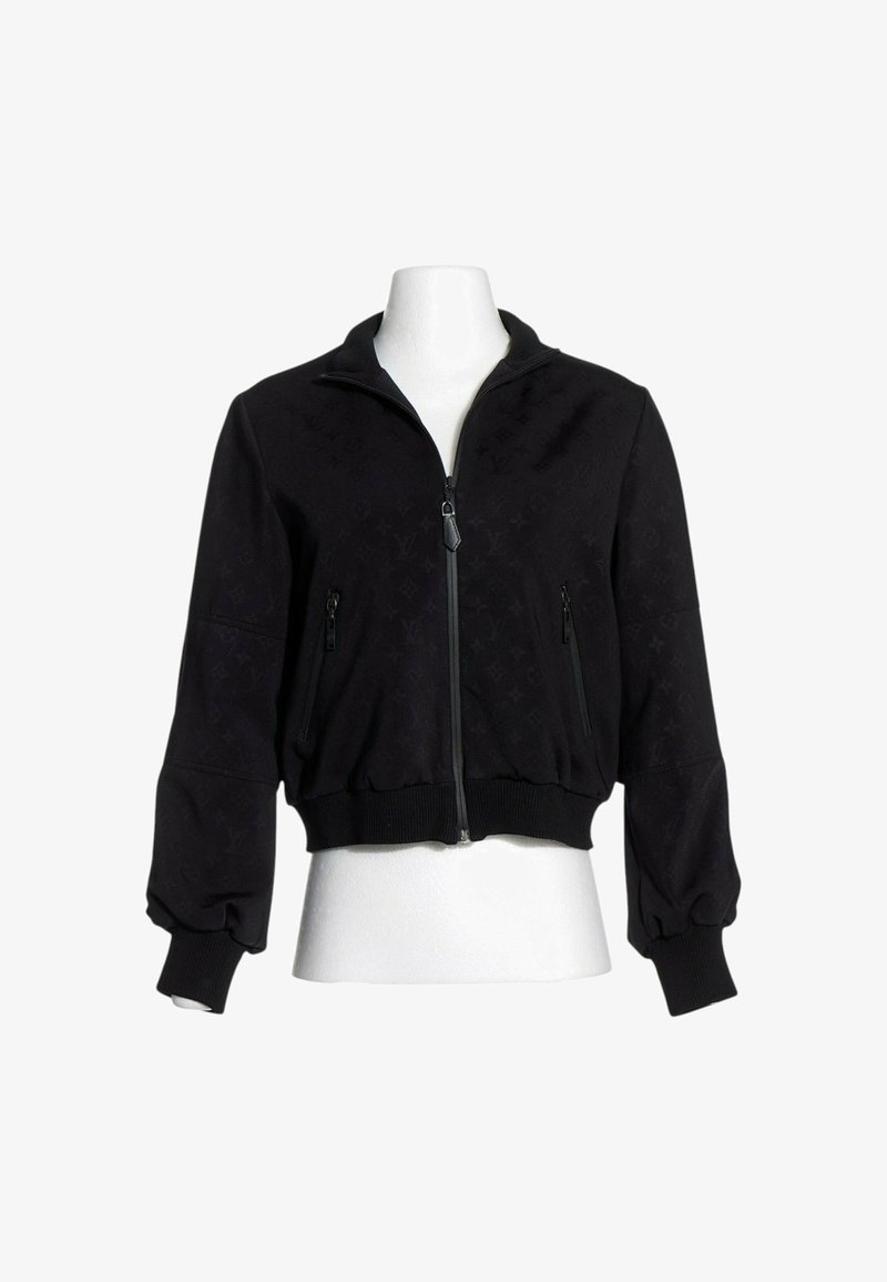 Veste noire zippée, manches longues, poignets et bas côtelés, avec deux poches avant zippées, présentée sur un torse de mannequin blanc.
