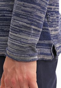 Lähellä kuva kädestä sinivalkoruudullisen pitkähihaisen paidan hihansuun vieressä, alhaalla näkyy tummat housut.