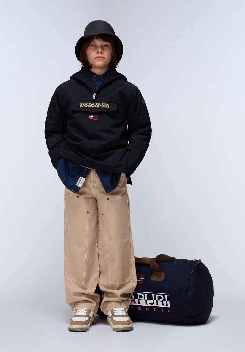 Zwarte capuchonjack met een logo patch, beige wijde broek en beige sneakers. Zwarte bucket hat en een marineblauwe sporttas met branding.