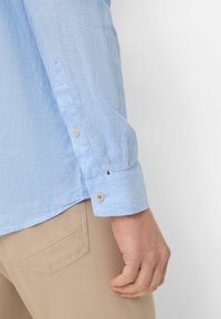 Lichtblauwe linnen shirt met een knoopsluiting aan de manchetten, met een geweven stof. Beige broek gedeeltelijk zichtbaar aan de onderlinkerkant.