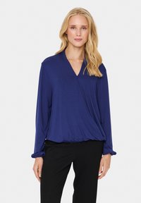 Maglietta a maniche lunghe blu navy con scollatura a V e fondo arricciato, realizzata in tessuto morbido e liscio. Indossata con pantaloni neri.