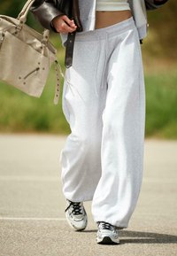 Osoba spacerująca na zewnątrz, nosząca jasnoszare dresy, białe i czarne trampki, trzymająca beżową torebkę oraz zakutą w ciemną kurtkę.